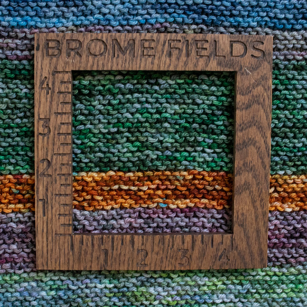 Close-up photo of a worsted yarn swatch in the garter stitch pattern, measured with a wooden 4-inch gauge ruler engraved with “Brome Fields.” The yarn is soft colorful, showing clear stitch definition and beautiful squishy texture.
