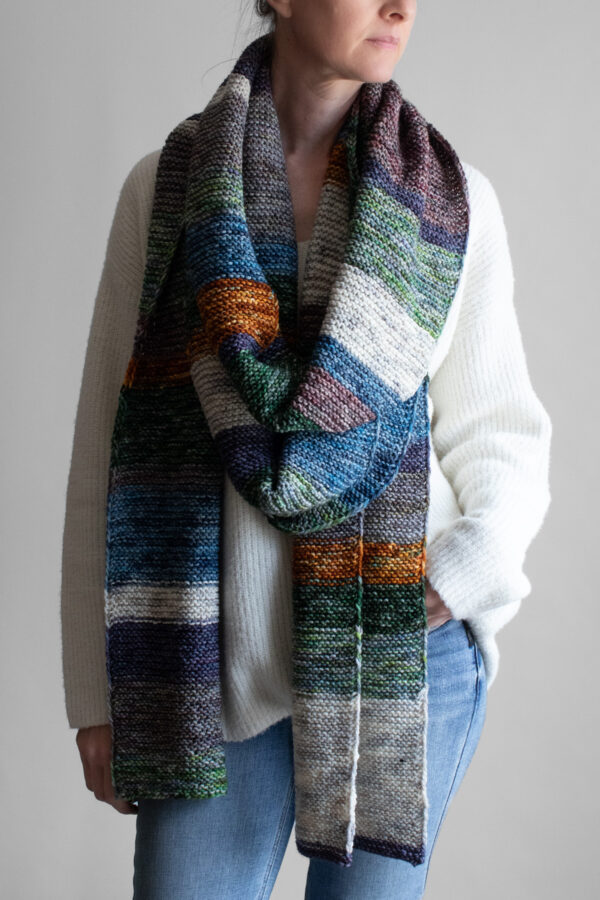 Woman wearing an extra long colorful striped garter stitch scarf in hand-dyed multicolor yarn, draped around her neck.