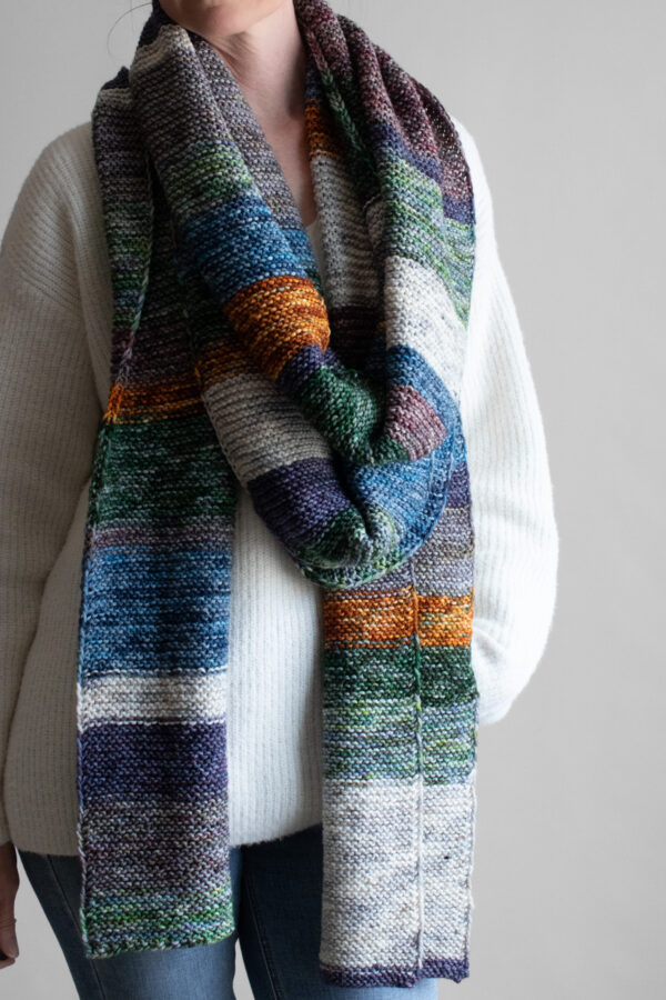 Woman wearing an extra long colorful striped garter stitch scarf in hand-dyed multicolor yarn, draped around her neck.