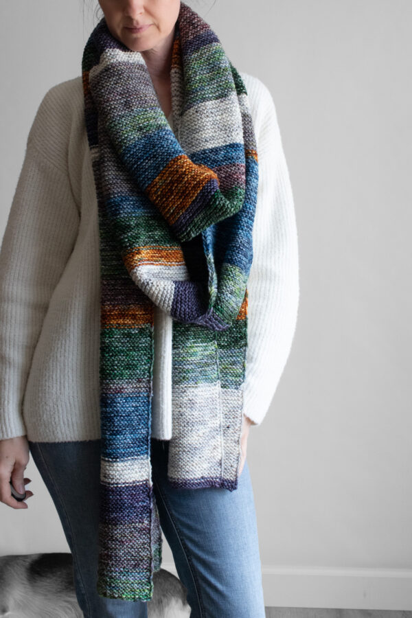 Woman wearing an extra long colorful striped garter stitch scarf in hand-dyed multicolor yarn, draped around her neck.