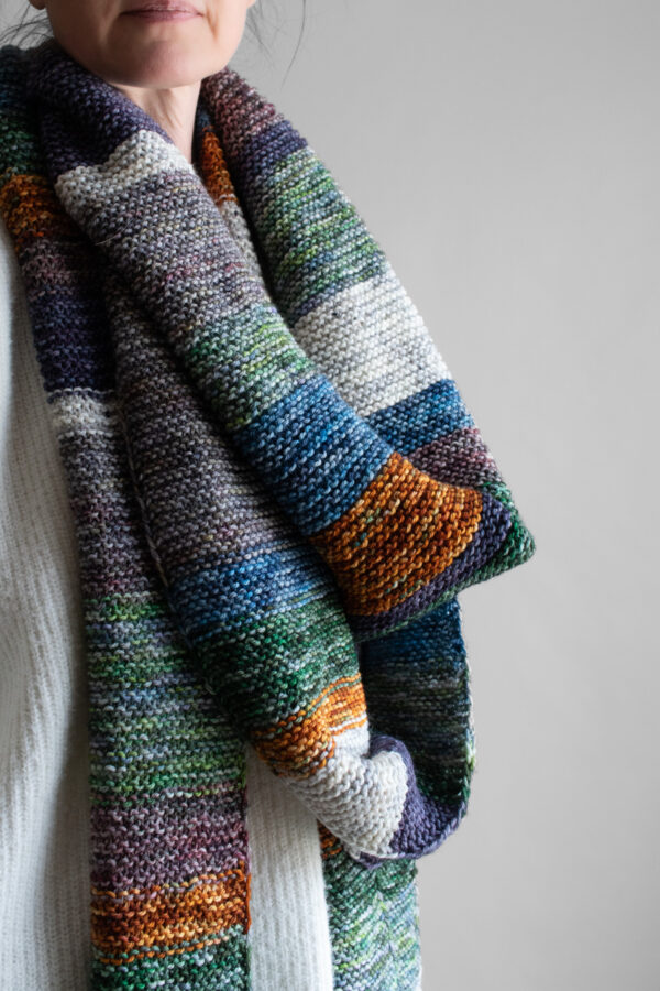 Woman wearing an extra long colorful striped garter stitch scarf in hand-dyed multicolor yarn, draped around her neck.