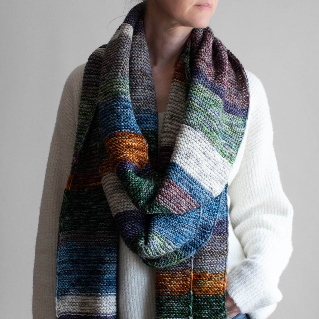 Woman wearing an extra long colorful striped garter stitch scarf in hand-dyed multicolor yarn, draped around her neck.