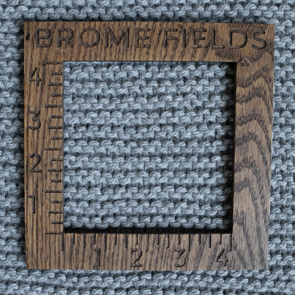 Close-up photo of a chunky yarn swatch in the garter stitch pattern, measured with a wooden 4-inch gauge ruler engraved with “Brome Fields.”