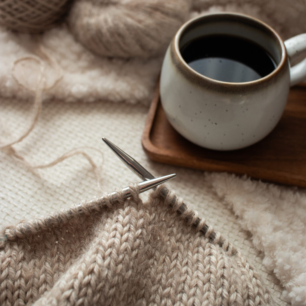 Cozy knitting flat lay with coffee cup, fuzzy ball of yarn, knitting needles with a new project WIP on a soft textile background