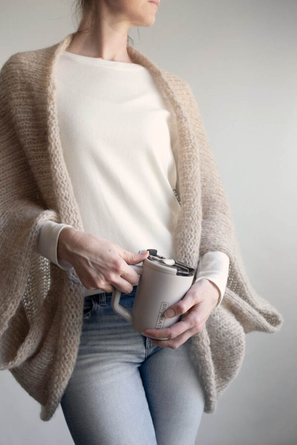 Woman wearing a loose knit, lightweight summer shrug over a cream tee and jeans holding a mug