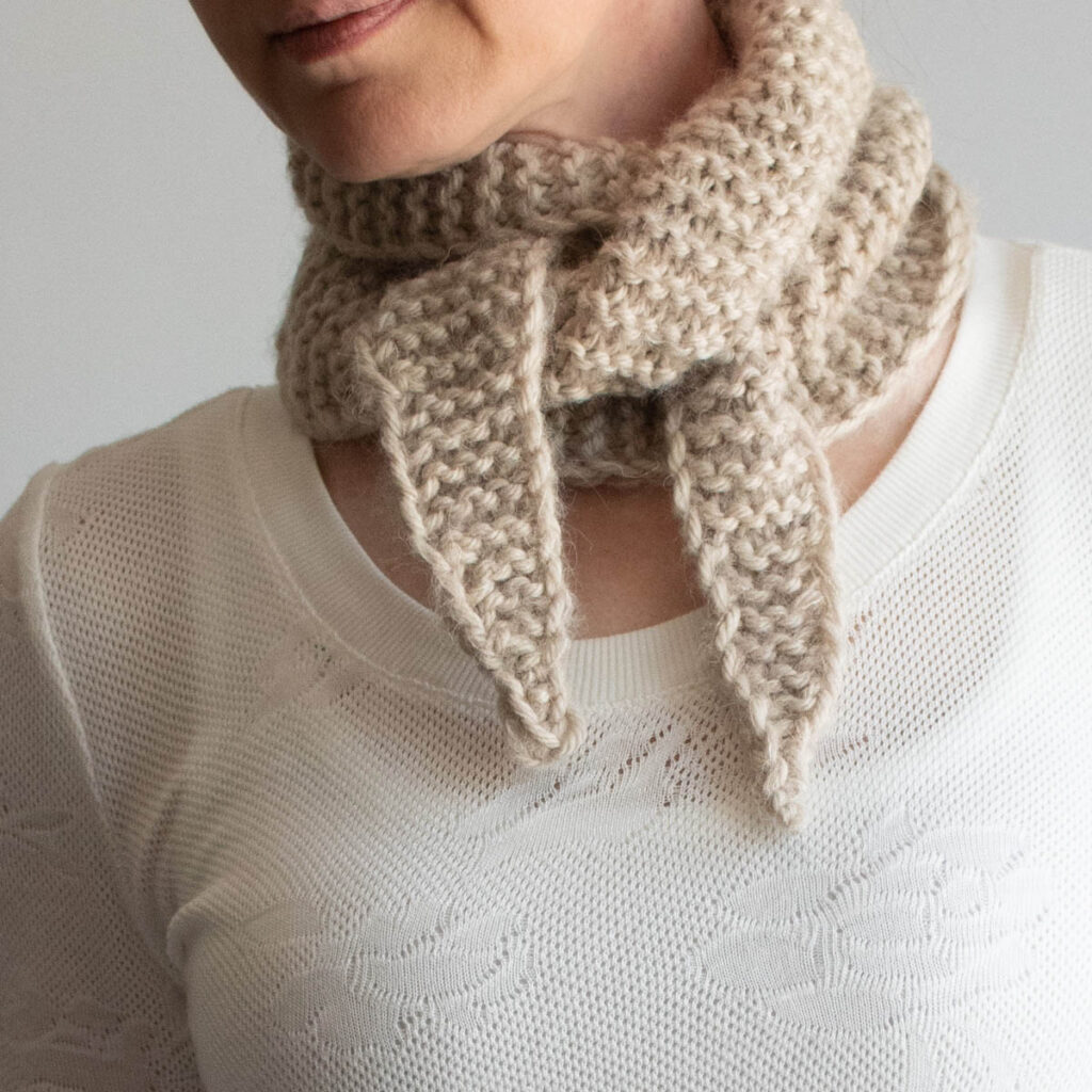 woman wearing a beige knit kerchief scarf wrapped and tucked around her neck, with two pointed tails hanging down, over a white sweater.