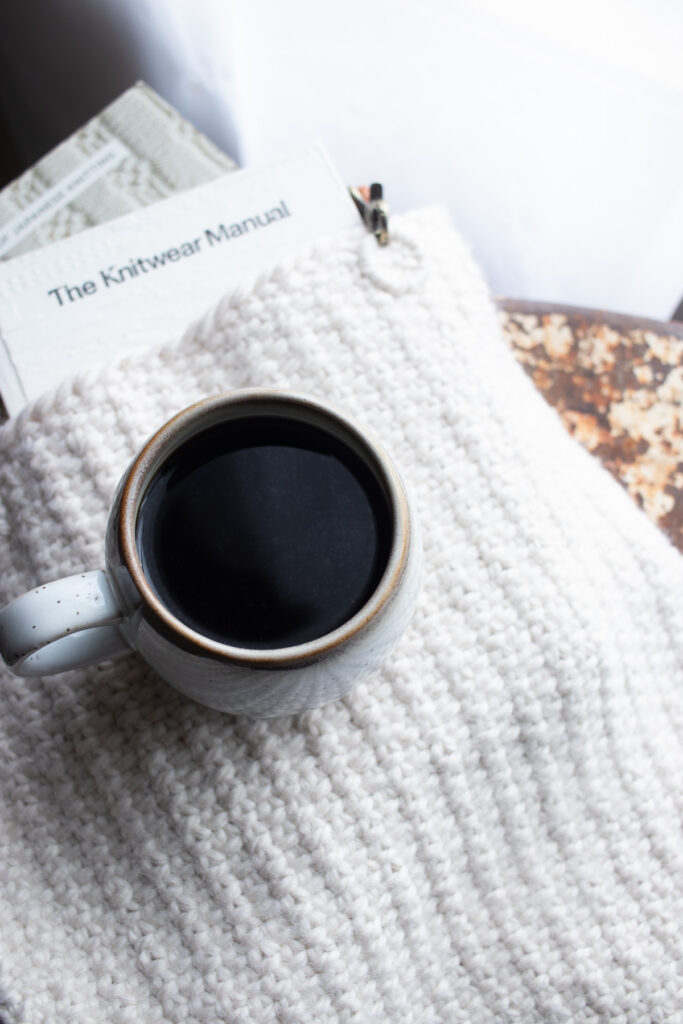 Pic of a linen stitch tote bag with books peeking out of the top sitting on a vintage metal chair with a cup of coffee on top