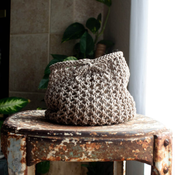 Close-up of a hand-knit beige mesh market bag with a crocheted drawstring tied in a bow, sitting on a rustic metal chair.