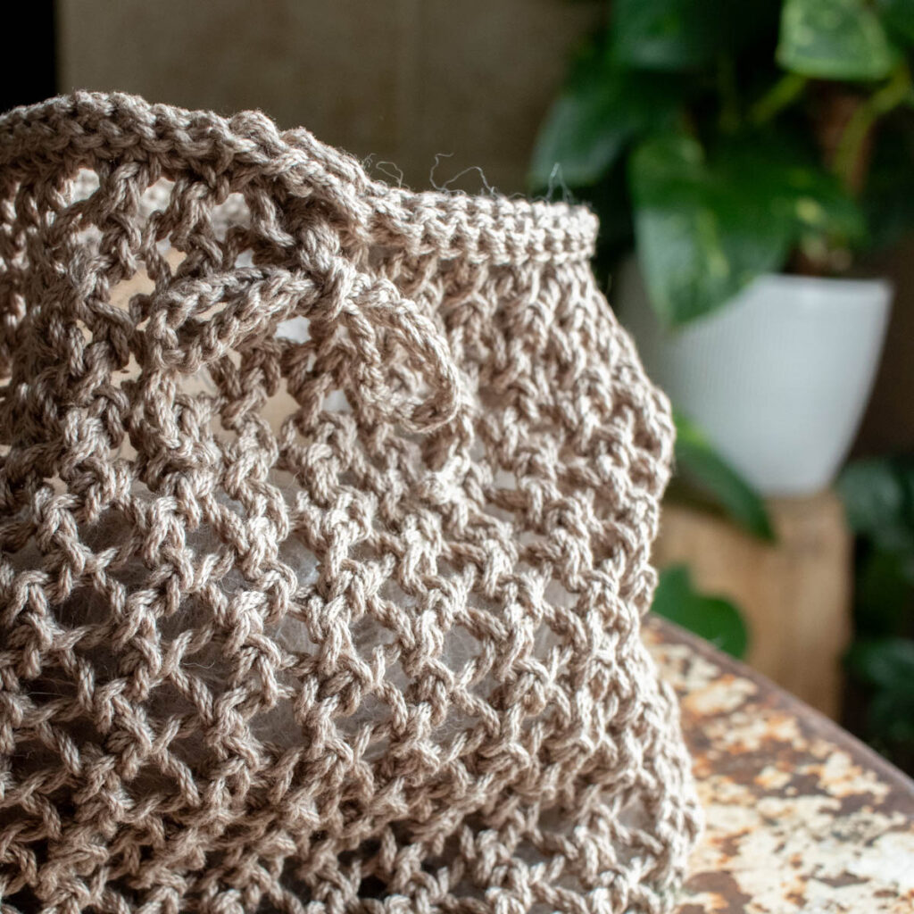 Open-weave knit market bag in natural beige yarn, styled with greenery in the background.
