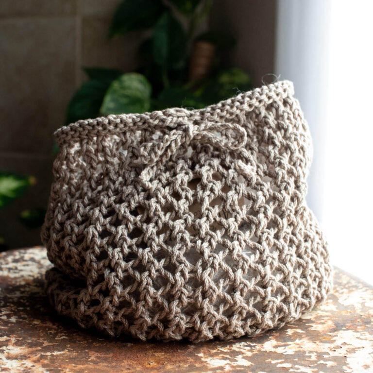 Hand-knit beige mesh market bag with a crocheted drawstring bow, photographed on a rustic table near a window.