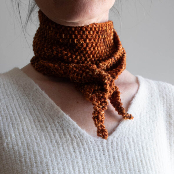 Woman wearing a rust-colored seed stitch skinny scarf kerchief-style around her neck.