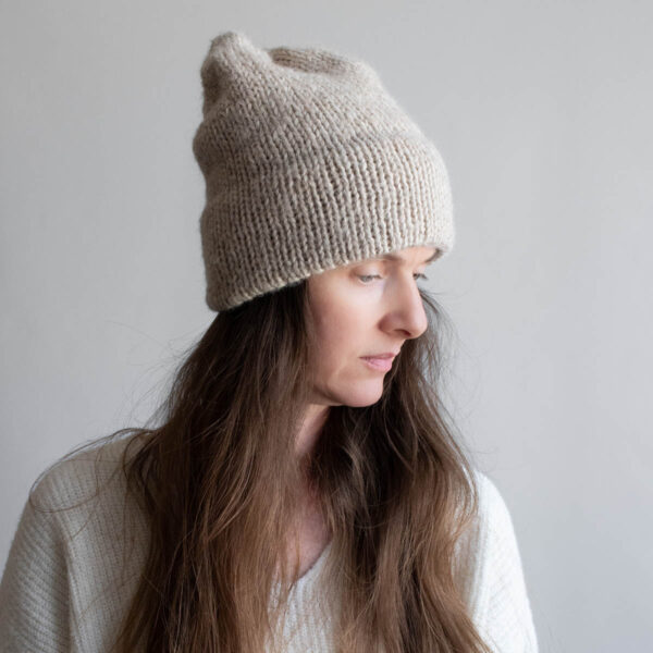 Woman with long brown hair wearing a slouchy oatmeal knit hat with a soft mohair halo, eyes downcast, photographed in profile against a soft gray background. White ribbed sweater visible at the shoulder.