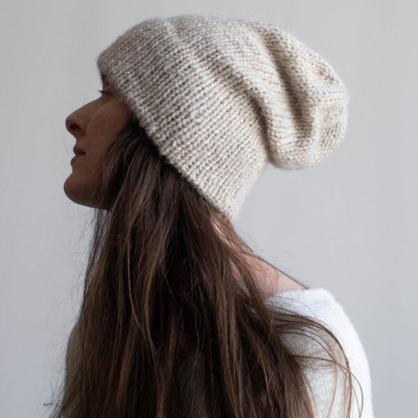 Woman with long brown hair wearing a slouchy oatmeal knit hat with a soft mohair halo, eyes downcast, photographed in profile against a soft gray background. White ribbed sweater visible at the shoulder.