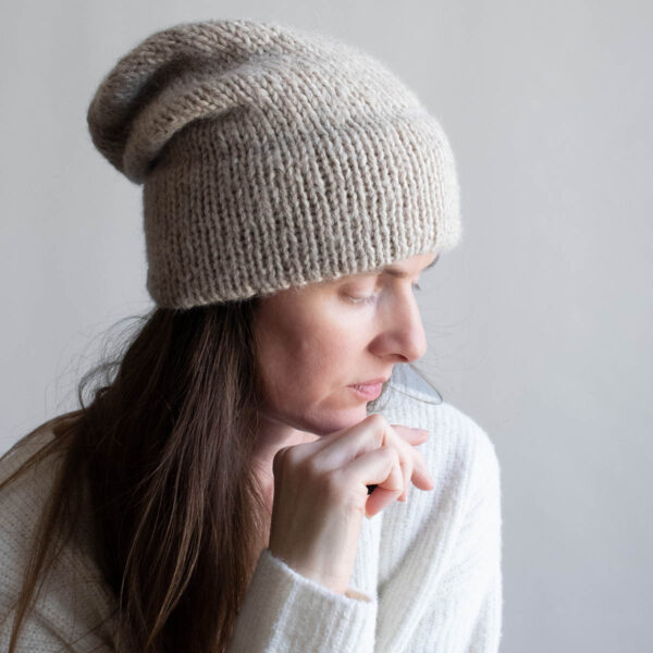 Woman with long brown hair wearing a slouchy oatmeal knit hat with a soft mohair halo, eyes downcast, photographed in profile against a soft gray background. White ribbed sweater visible at the shoulder.