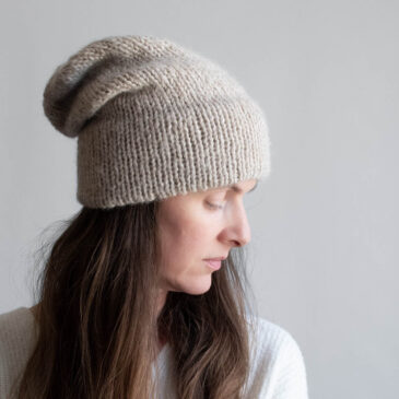 Woman with long brown hair wearing a slouchy oatmeal knit hat with a soft mohair halo, eyes downcast, photographed in profile against a soft gray background. White ribbed sweater visible at the shoulder.