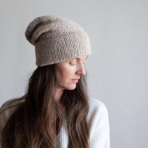 Woman with long brown hair wearing a slouchy oatmeal knit hat with a soft mohair halo, eyes downcast, photographed in profile against a soft gray background. White ribbed sweater visible at the shoulder.