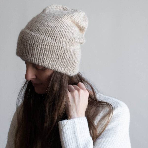 Woman with long brown hair wearing a slouchy oatmeal knit hat with a soft mohair halo, eyes downcast, photographed in profile against a soft gray background. White ribbed sweater visible at the shoulder.