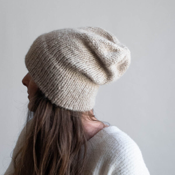 Woman with long brown hair wearing a slouchy oatmeal knit hat with a soft mohair halo, eyes downcast, photographed in profile against a soft gray background. White ribbed sweater visible at the shoulder.