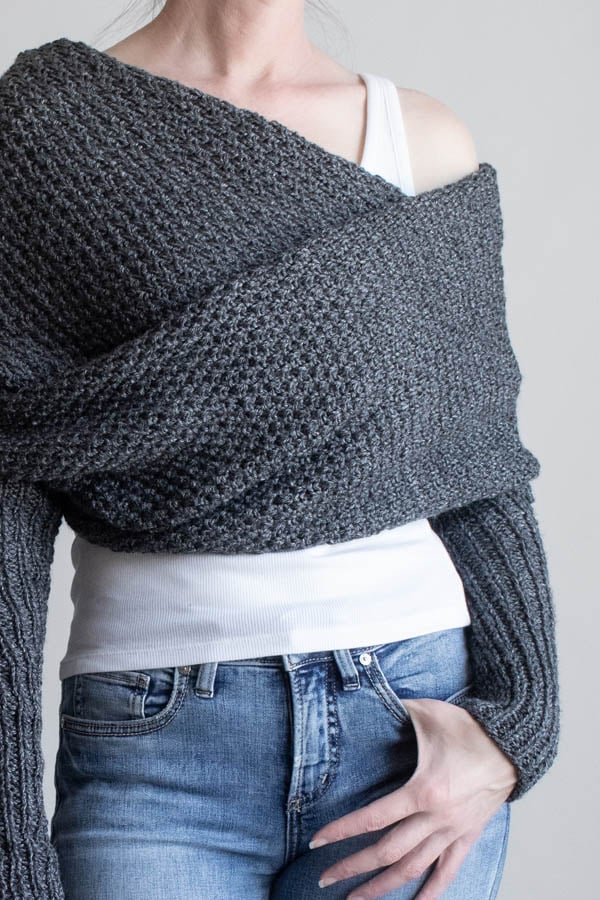 Full front view of a woman wearing a charcoal moss stitch sweater scarf wrapped across her shoulders, with long ribbed sleeves, over a white tank and jeans