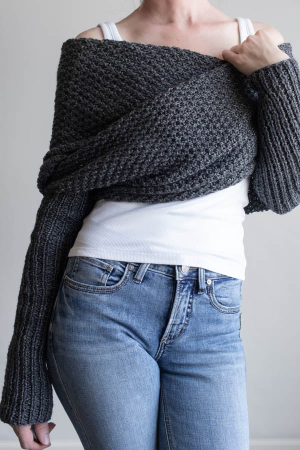 Full front view of a woman wearing a charcoal moss stitch sweater scarf wrapped across her shoulders, with long ribbed sleeves, over a white tank and jeans
