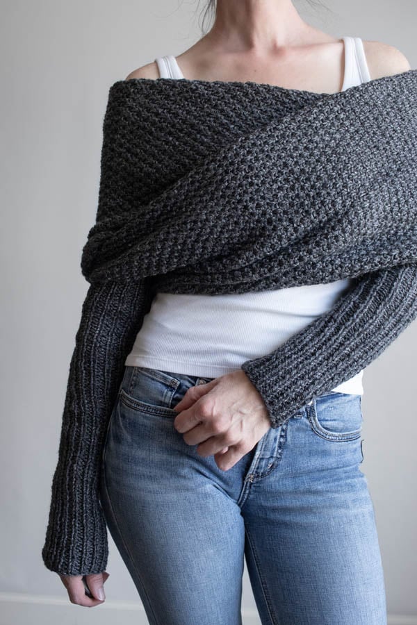 Full front view of a woman wearing a charcoal moss stitch sweater scarf wrapped across her shoulders, with long ribbed sleeves, over a white tank and jeans