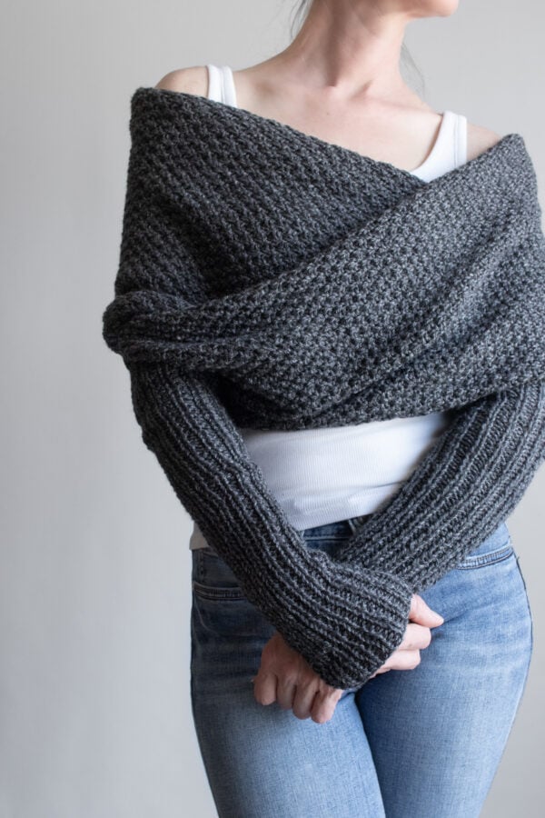 Full front view of a woman wearing a charcoal moss stitch sweater scarf wrapped across her shoulders, with long ribbed sleeves, over a white tank and jeans
