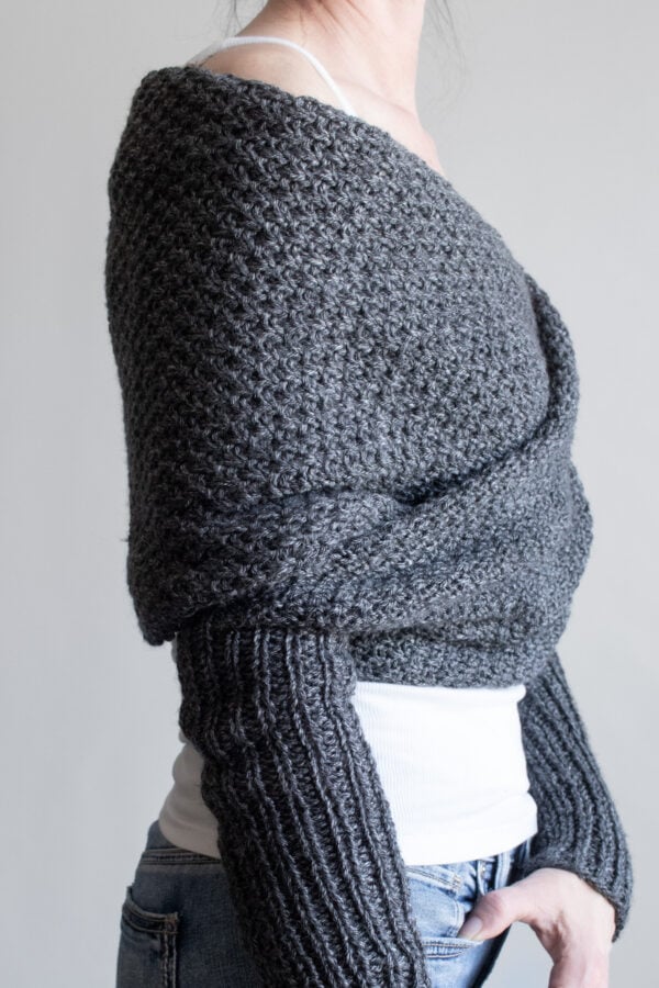 Side view of a woman wearing a charcoal moss stitch sweater scarf wrapped across her shoulders, with long ribbed sleeves, over a white tank and jeans