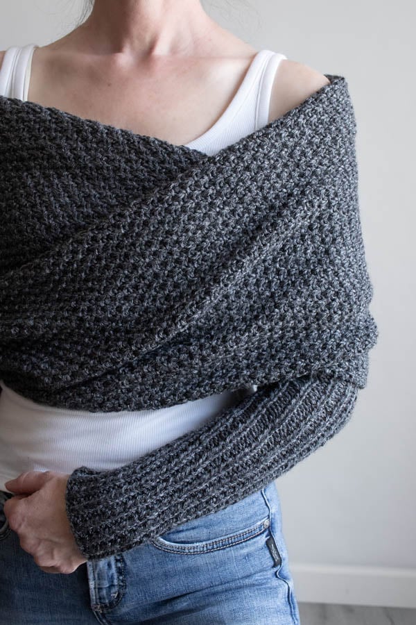 Woman wearing a charcoal moss stitch sweater scarf wrapped across her shoulders, with long ribbed sleeves, over a white tank and jeans