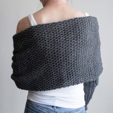 Back view of a woman wearing a charcoal moss stitch sweater scarf wrapped across her shoulders, with long ribbed sleeves, over a white tank and jeans
