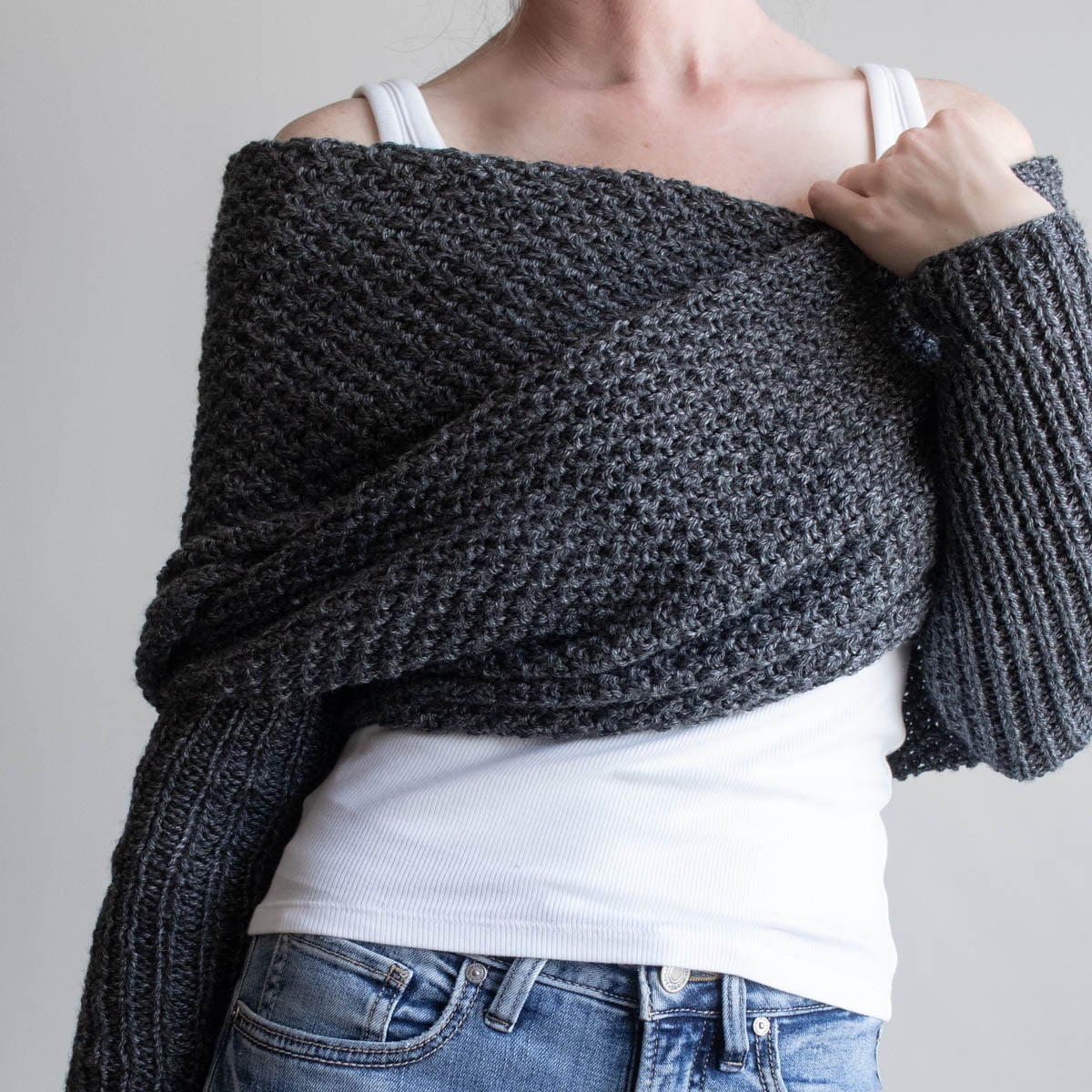 Woman wearing a charcoal moss stitch sweater scarf wrapped across her shoulders, with long ribbed sleeves, over a white tank and jeans